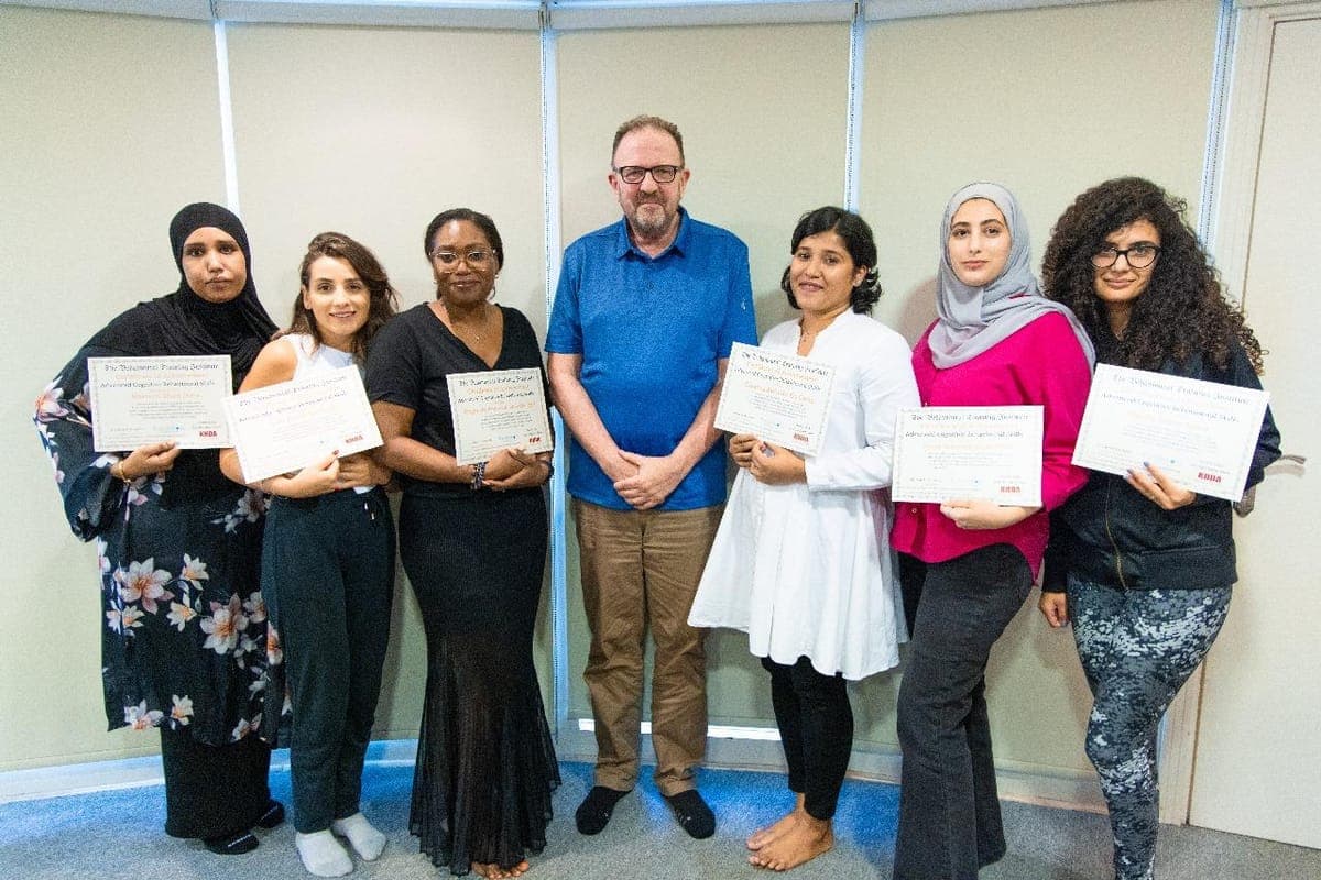 Six diverse graduates holding CBT Skills certificates with Dr. John Crimmins at a professional training venue